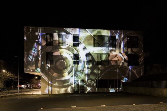 Eine nächtliche Außenansicht eines modernen Gebäudes, dessen Fassade mit einer großflächigen, abstrakten Videoprojektion aus geometrischen Formen, Kreisen und Lichtreflexen in warmen Braun-, Gold- und Grüntönen beleuchtet wird. Die Projektion überlagert die Fenster und architektonischen Strukturen des Gebäudes. Im Vordergrund befindet sich ein leerer, gepflasterter Platz, während Straßenlaternen und Bäume die nächtliche Umgebung ergänzen.